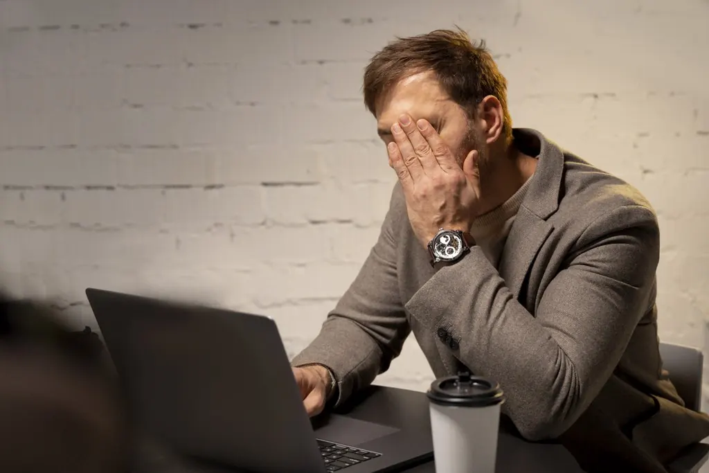 A frustrated man covering his face in front of a laptop